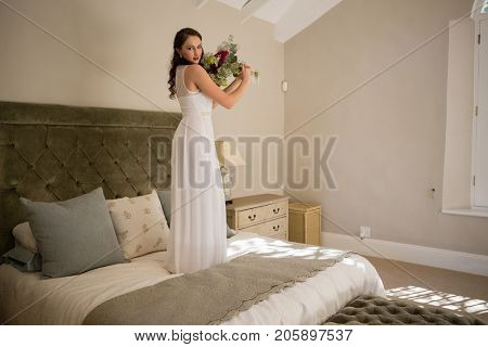 Portrait of beautiful bride holding bouquet while standing on bed at home