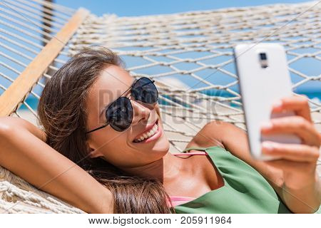 Vacation woman using smart phone taking smartphone selfie. Girl relaxing on beach hammock sun tanning in sunglasses smiling using cellphone to take pictures and text sms messages.