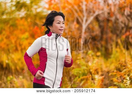 Active woman running and jogging. Middle aged Asian mature female jogger outdoor living healthy lifestyle in beautiful autumn city park in colorful fall foliage. Asian Chinese lady in her fifties.