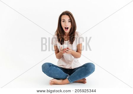 Shocked happy brunette woman holding smartphone in hands and looking at the camera over white background