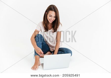 Pleased brunette woman using laptop computer and sitting on the floor over white background