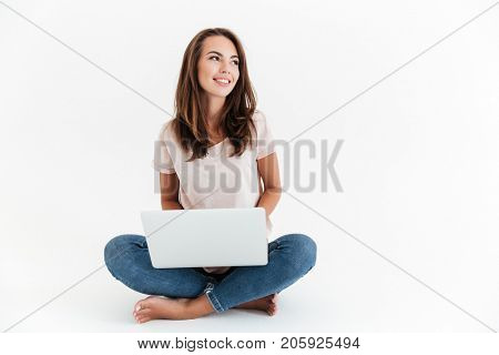 Smiling brunette woman holding laptop computer and looking away while sitting on the floor over white background