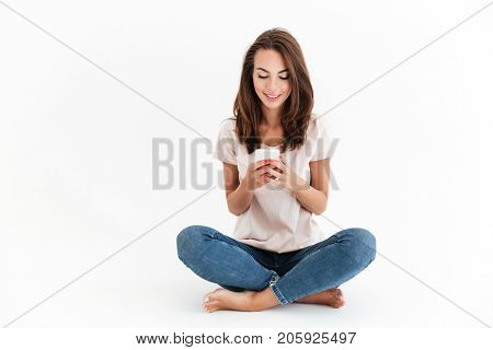 Smiling brunette woman sitting on the floor and using smartphone over white background