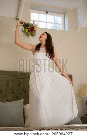 Low angle view of beautiful bride holding bouquet singing while standing on bed at home