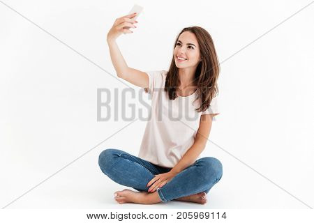 Smiling brunette woman sitting on the floor and making selfie on smartphone over white background