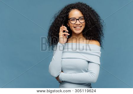 Indoor Shot Of Positive Lovely Woman Has Telephone Conversation, Keeps Mobile Phone Near Ear, Focuse