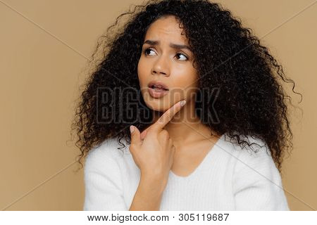 Puzzled Female Model Keeps Fore Finger On Chin, Looks Thoughtfully Aside, Has Frustrated Facial Expr