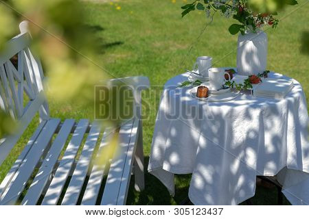 White Served Table With Coffee, Caneles And Flowers Next To White Bench In The Garden. Enjoying Sunn