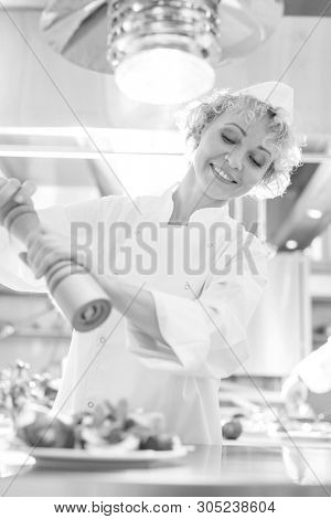 Black and White photo of Smiling mature chef using peppermill on salad in plate at restaurant