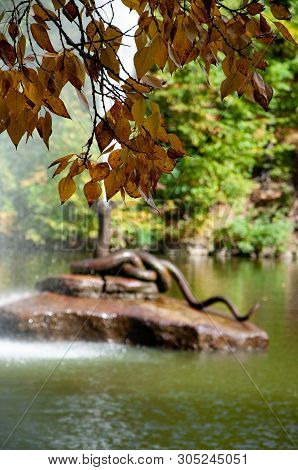 Bronze Snake Sculpture Fountain On Rock Island In Lake. Focus On Branch With Brown Autumn Leaves On 