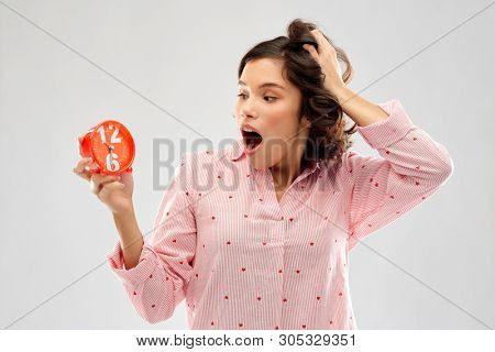 time, emotions and bedtime concept - shocked young woman in pajama with alarm clock showing half past six over grey background