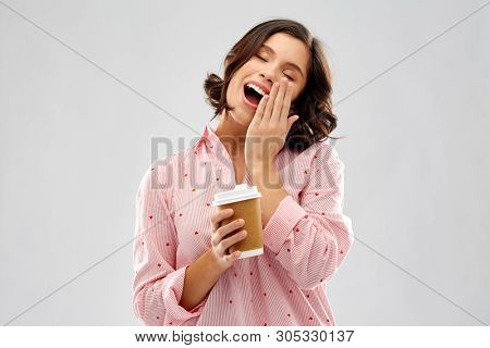 people and bedtime concept - happy sleepy yawning young woman in pajama with takeaway paper cup of coffee over grey background