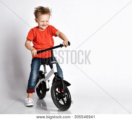 Happy Kid Boy In Orange T-shirt And Blue Jeans Tries To Ride His New Black And White Bicycle Without