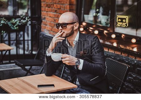 Pensive Modern Man Is Smoking A Cigarette Outside Of Coffeshop While Drinking His Cup Of Coffee.