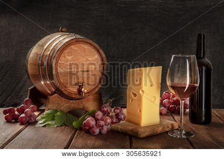 A Glass Of Red Wine With A Bottle, A Wine Barrel, A Large Piece Of Cheesse, Grapes, And Vine Leaves,