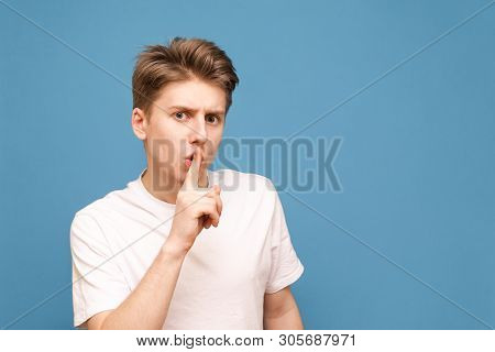 Guy In A White T-shirt Put His Finger To The Lips And Shows A Sign Of Silence, Isolated On A Blue Ba