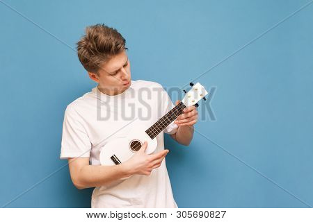Concentrated Young Man In A White T-shirt Stands On A Blue Background With A Ukulele In Hands And Lo