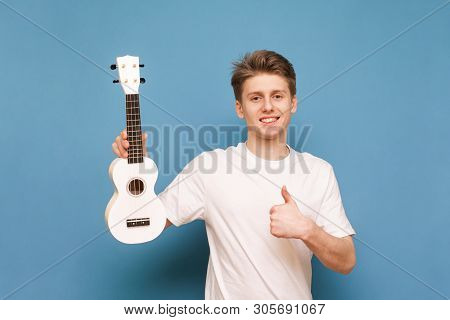 Happy Young Man Standing With Ukulele In His Hands On A Blue Background, Showing A Thumbs Up, Lookin