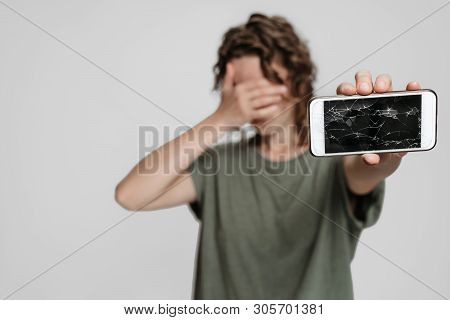 Frustrated Curly Woman Covers Face With Palm, Showing Her Broken Smartphone, Isolated On Gray Backgr