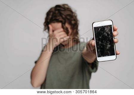 Upset Curly Woman Covers Face With Palm, Showing Her Broken Smartphone With Cracked Screen Protectio