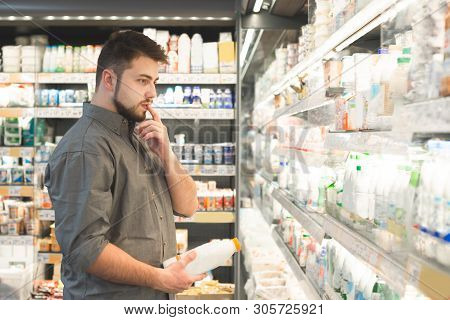 Portrait Of A Pensive Man Standing In The Supermarket Dairy Department, Looks At Shelves With Bottle