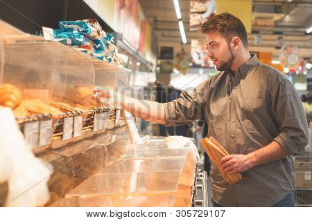 Man Buys Food In The Bread Department Of The Supermarket.man In A Shirt Holds A Paper Bag In His Han