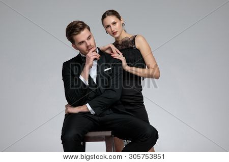 Seated groom looking to the side and thinking with his hand on his chin while his girlfriend leans on his shoulder on gray studio background