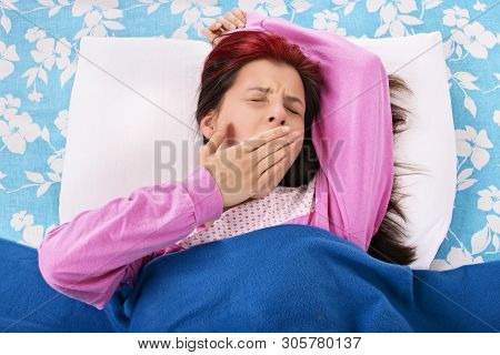 Young Beautiful Girl Lying On A Pillow Under A Blanket Yawning, Isolated On A White Background.