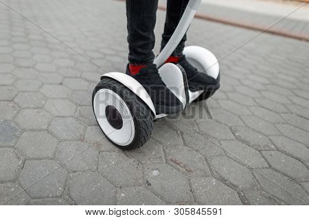 Closeup Of A Man's Feet On A Modern Hoverboard. Stylish Guy In Jeans In Sneakers Standing On A White