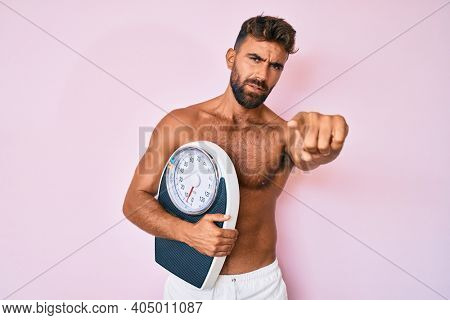 Young hispanic man standing shirtless holding weighing machine pointing with finger to the camera and to you, confident gesture looking serious 