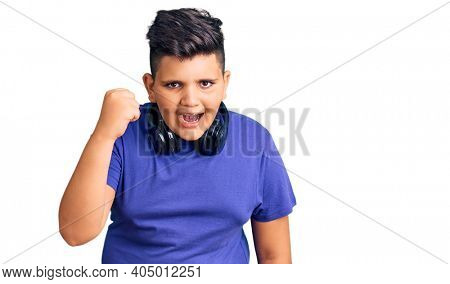 Little boy kid listening to music wearing headphones angry and mad raising fist frustrated and furious while shouting with anger. rage and aggressive concept. 
