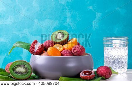 Lychee, Kiwi And Kumquat In A Gray Ceramic Bowl And A Glass Of Water Against The Turquoise Backgroun