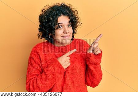 Young hispanic woman with curly hair wearing casual winter sweater pointing aside worried and nervous with both hands, concerned and surprised expression 