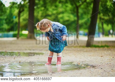Child Wearing Red Rain Boots And Jumping In Puddle On A Summer Day. Adorable Toddler Girl Having Fun
