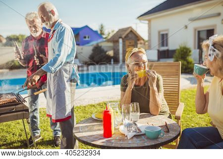Senior Women Having Fun Drinking Coffee And Relaxing While Men Are Grilling Meat And Drinking Beer I