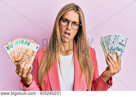 Young blonde woman wearing business style holding dollars and euros banknotes in shock face, looking skeptical and sarcastic, surprised with open mouth 
