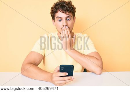 Young caucasian man with curly hair using smartphone sitting on the table covering mouth with hand, shocked and afraid for mistake. surprised expression 