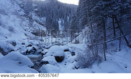 A Clear Mountain River Runs Through A Snowy Gorge. There Is A Metal Bridge Covered With Snow. Tall S