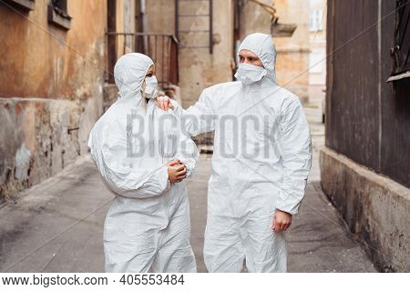 Doctors In Protective Suits And Protective Masks Stand On The Street With Serious Faces. Walking Alo