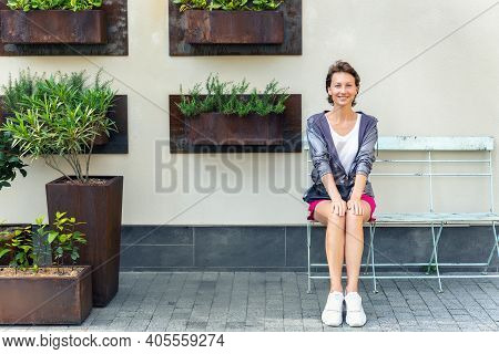 Young Adult Beautiful Caucasian Woman Sitting On Metal Vintage Bench Chair Against Wall With Many Mo