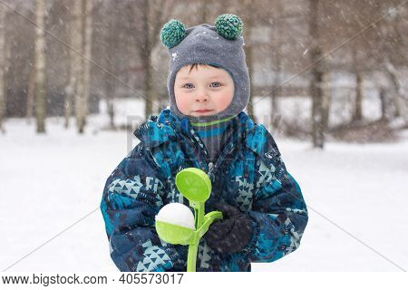 A Boy In A Winter Overalls Stands On The Street, In The Hands Of A Snowball. The Child Makes Snowbal