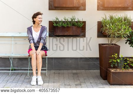 Young Adult Beautiful Caucasian Woman Sitting On Metal Vintage Bench Chair Against Wall With Many Mo