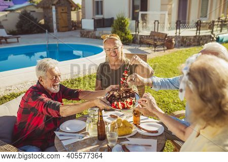 Group Of Cheerful Senior Friends Eating Lunch In The Backyard By The Pool, Gathered Around The Table