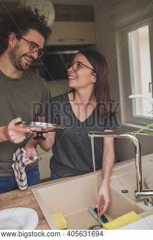Beautiful Young Couple In Love Doing The Washing Up Together After Lunch; Woman Washing The Dishes W