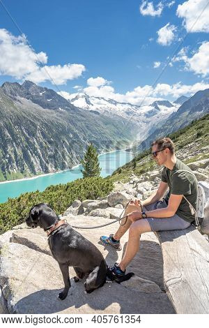 Zillertal, Austria - Aug 7, 2020: Male Hiker Rest On Bench With His Black Dog On Way To Olpererhutte