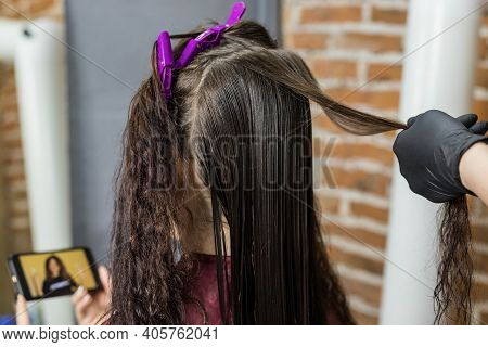 A Young Woman Watches A Video On Her Phone While Sitting In A Hair Salon For A Keratin Hair Straight