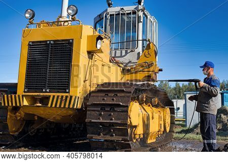 Car Wash Of Heavy Machinery And Tractors. A Man In Work Clothes And A Medical Mask Washes A Yellow C