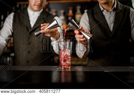 Bartenders Hold Steel Jiggers And Pour Drink Into Glass With Ice Cubes