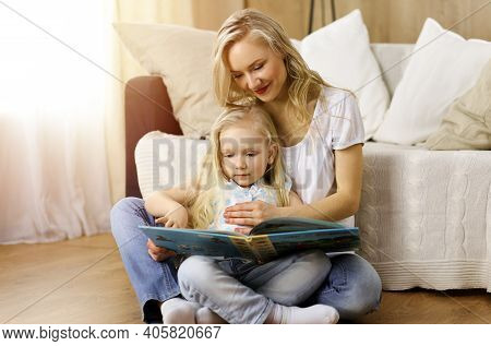Happy Family. Blonde Young Mother Reading A Book To Her Cute Daughter While Sitting At Wooden Floor 