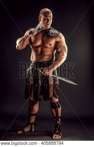 Severe Barbarian In Leather Costume With Two Swords. Portrait Of Balded Muscular Gladiator. Studio S